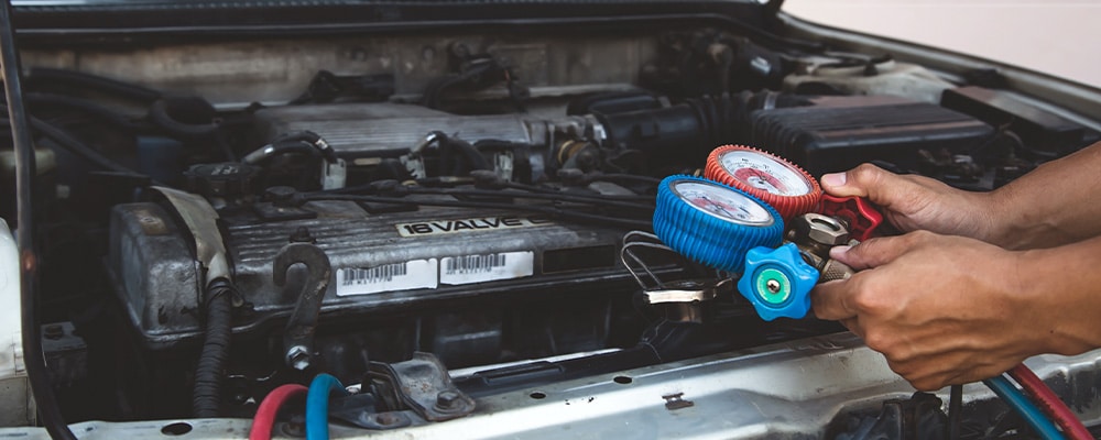 My car uses R1234yf refrigerant - Can I replace it with R134a? With Durand Automotive. Image of mechanic’s hands holding auto air conditioning machine over open car engine in shop.